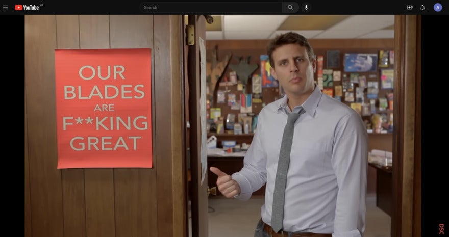 Dollar Shave Club video still of man pointing to a poster that says "Our Blades Are F**king Great"