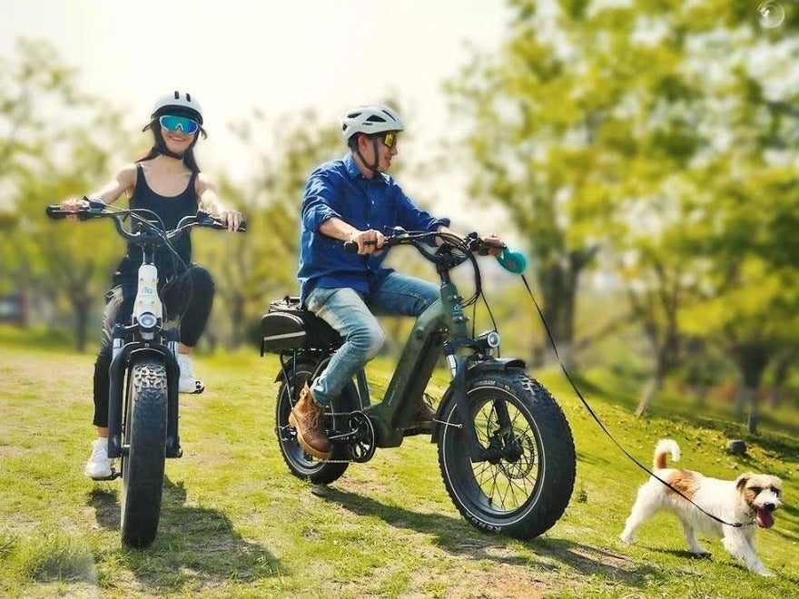 A man and a woman on electric bikes, riding across a field with trees in the background. The man is holding a dog on a lead.